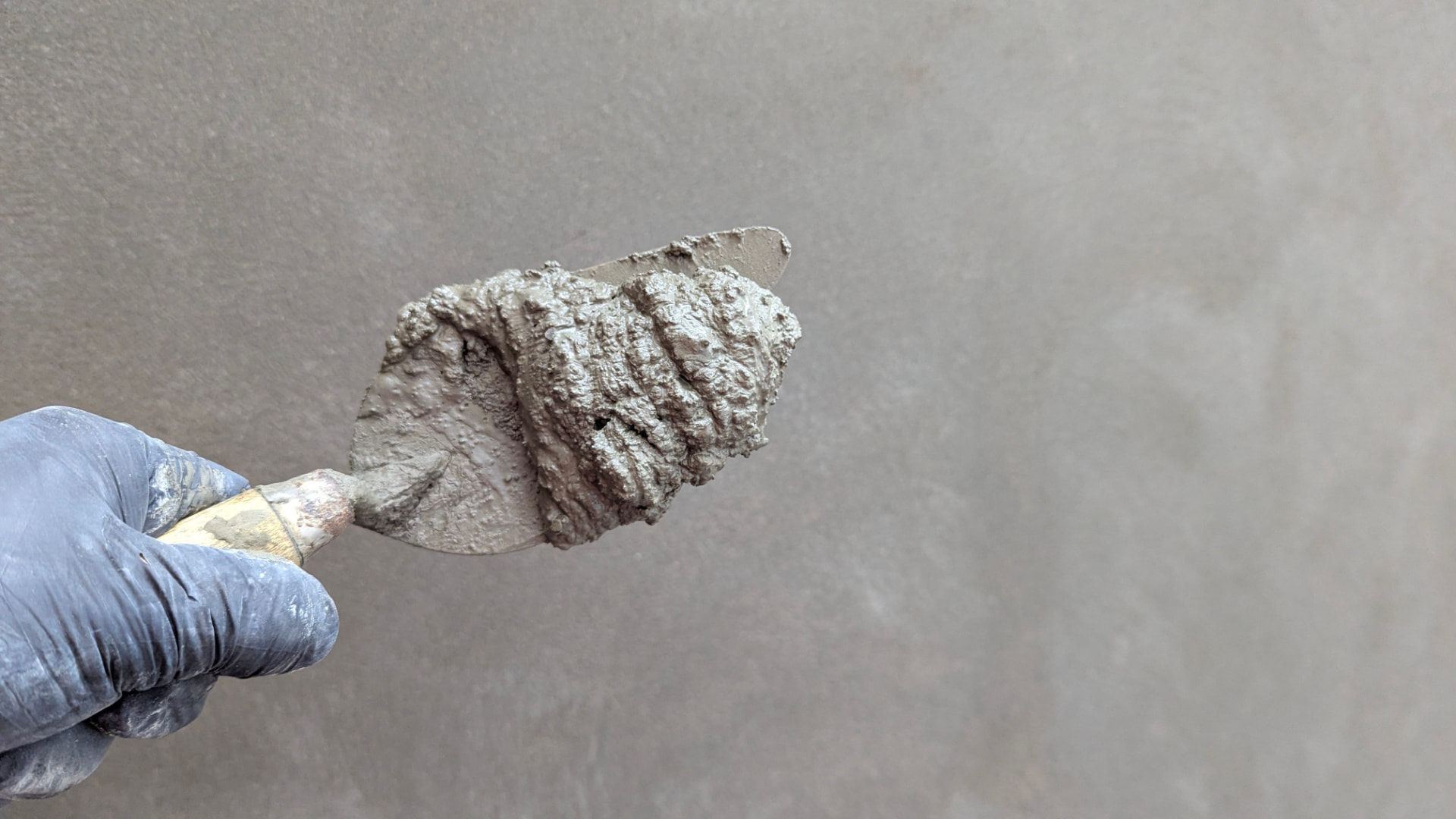 Gloved hand holding a trowel loaded with concrete repair mortar against a gray wall